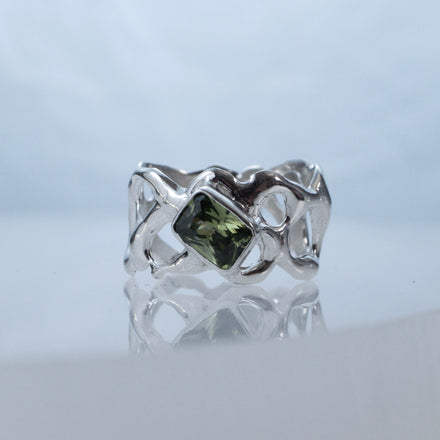 Silver sculptural and fluid ring with negative spaces and a green gemstone on a reflective surface