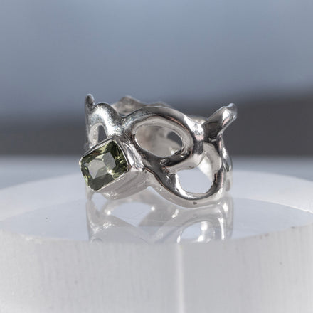 close up of a sculptural silver ring with flowing intricate negative spaces and a green gemstone on a reflective surface