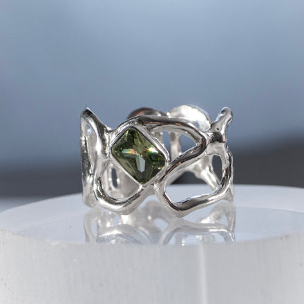 close up of a sculptural silver ring with flowing intricate negative spaces and a green gemstone on a reflective surface