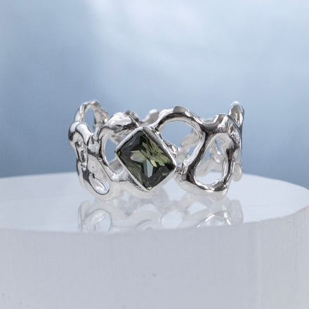 close up of a sculptural silver ring with flowing intricate negative spaces and a green gemstone on a reflective surface and light blue background