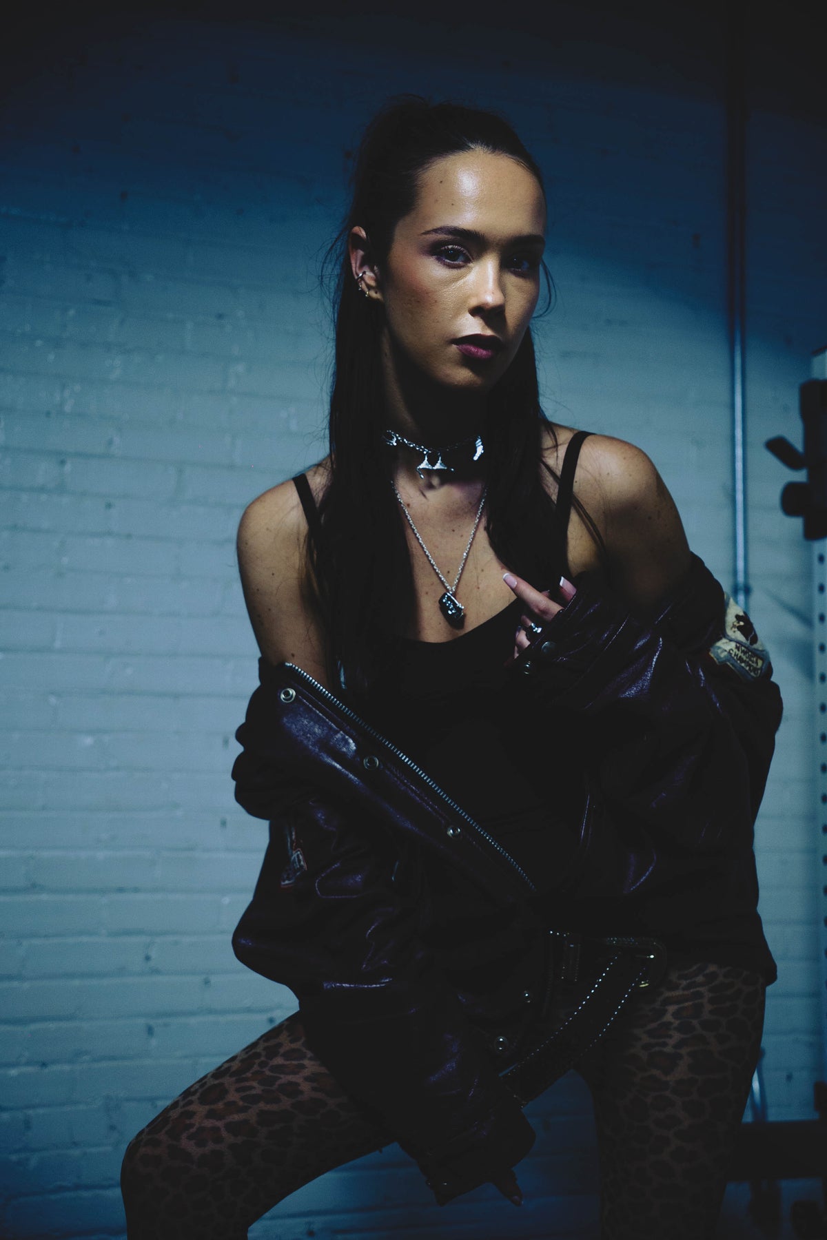Woman in dark outfit against a blue brick wall, wearing a leather bomber, leopard tights, sterling silver charm necklaces