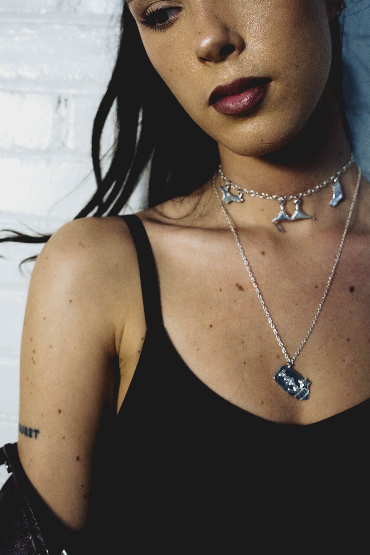 Close-up of a person wearing sterling silver necklaces with pendant charms against a white brick wall.