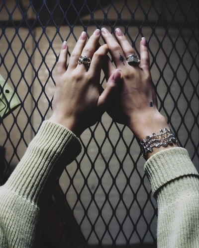 two hands outstretched with beige sweater sleeves in front of industrial wire fence. wearing statement silver rings and bracelet
