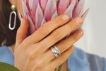 close up of a female hand holding a spiky flower, while display the silver blob ring, stacked on top of a similar ring that is thicker, all on her ring finger